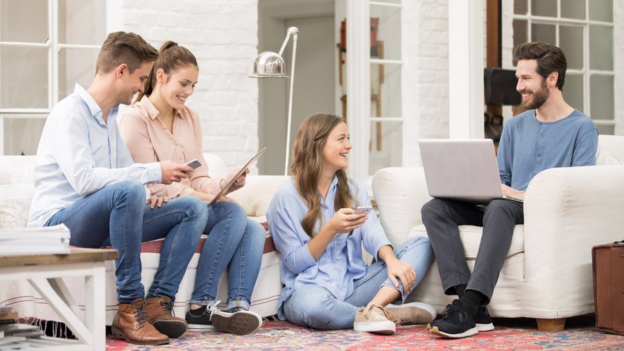 Vier junge Erwachsene sitzen gemeinsam auf einem Sofa, arbeiten an Laptop und Tablet und unterhalten sich