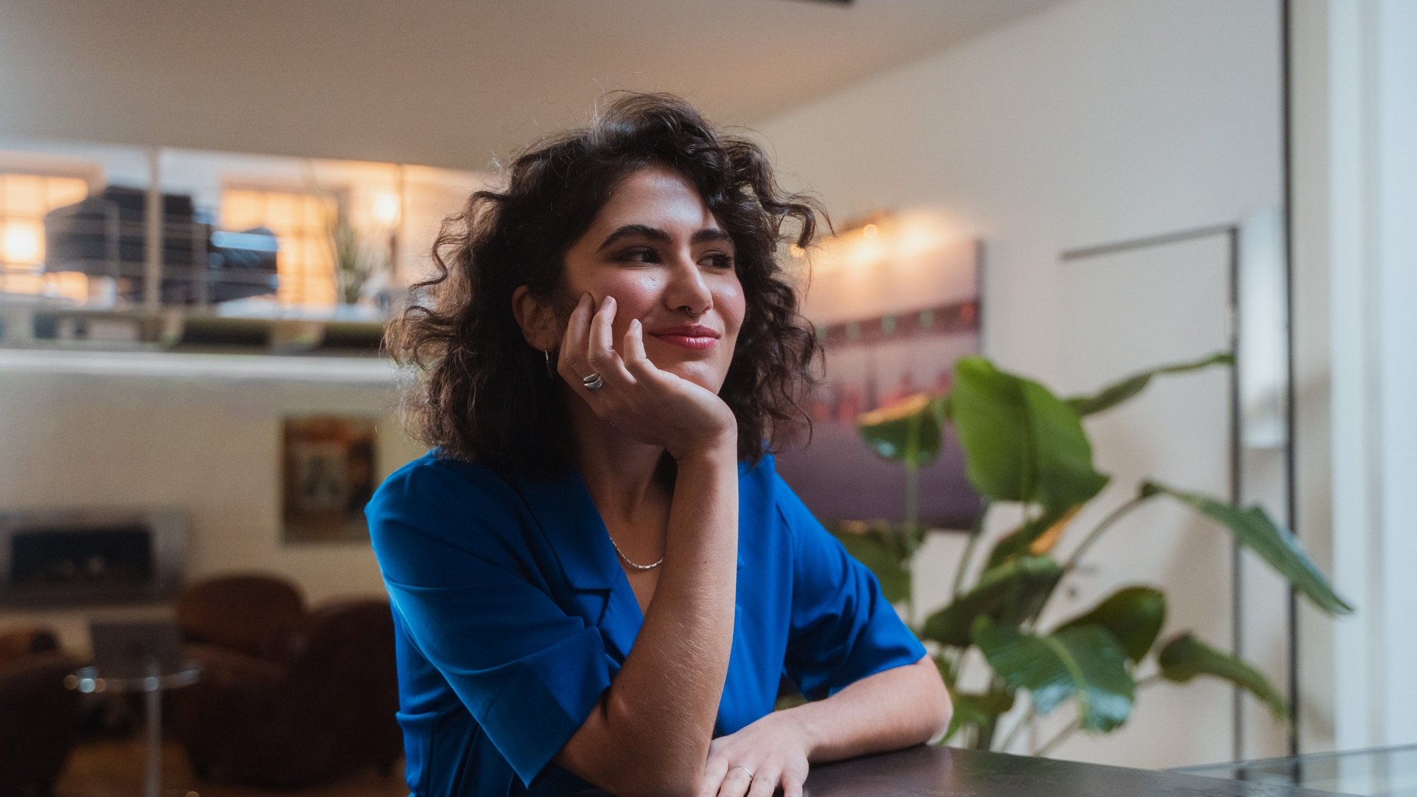 Frau mit blauer Bluse sitzt an einem Tisch und blickt nachdenklich in die Ferne.