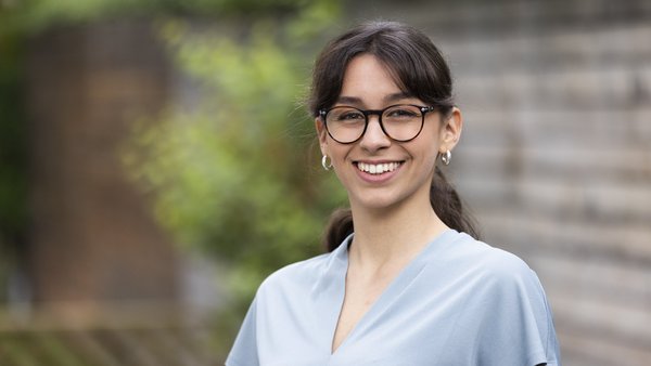Junge Studentin mit Brille Junge Frau mit Brille lächelt in die Kamera, steht im Freien vor einem grünen Hintergrund.